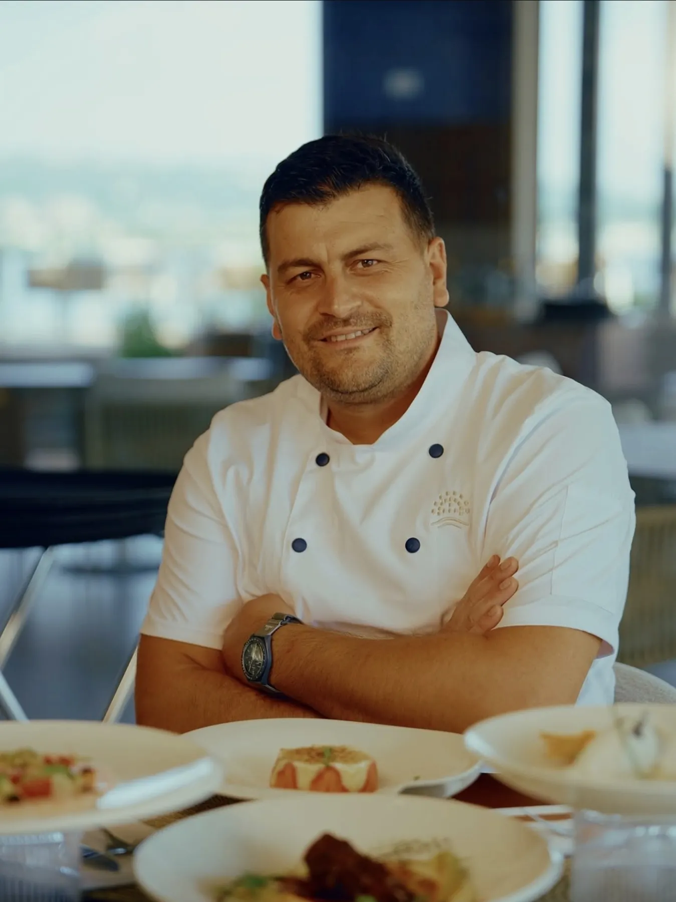 Portret Miloša Ristića u kuvarskoj bluzi, za stolom sa nekoliko serviranih jela.